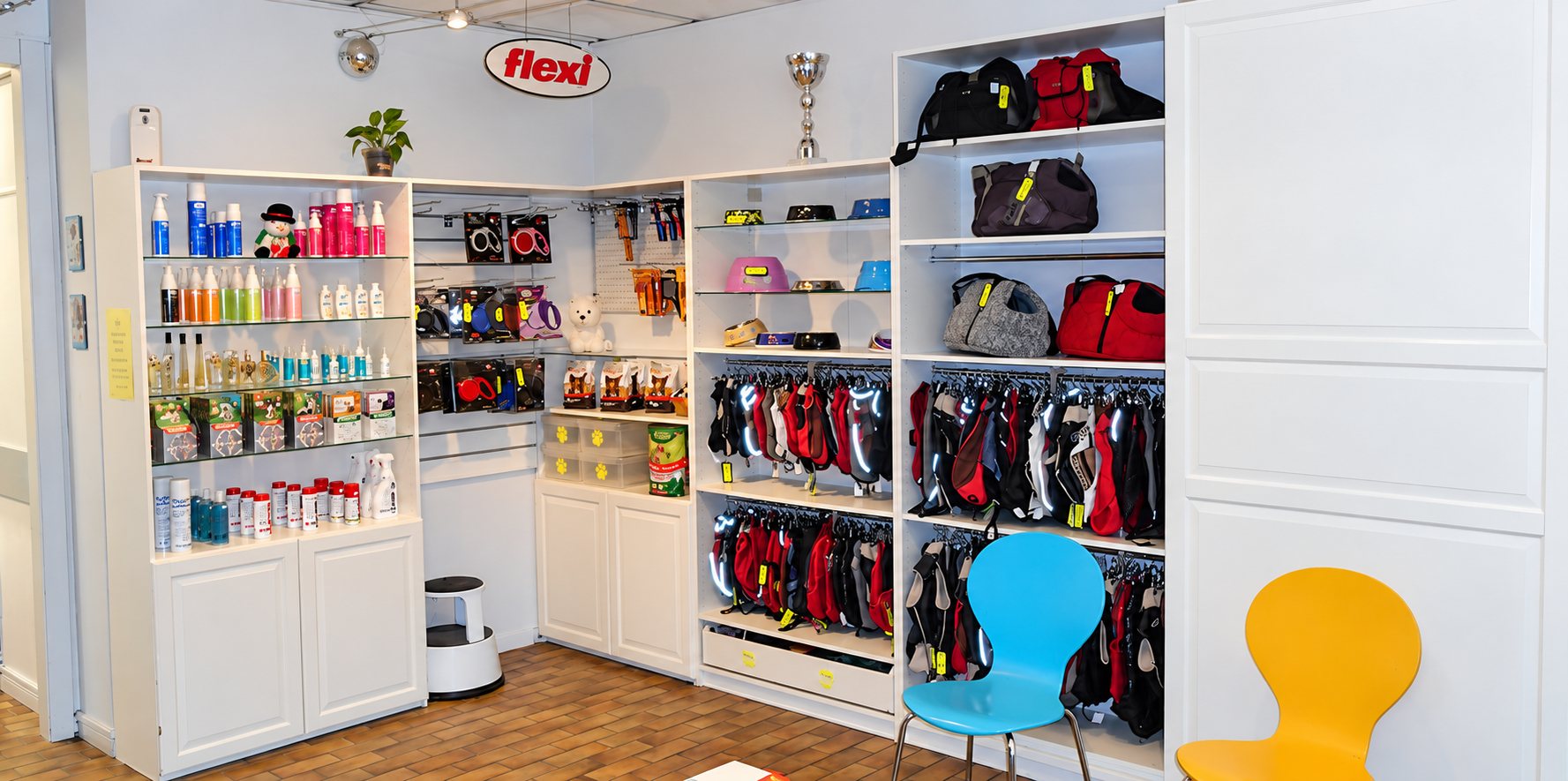Intérieur d'une boutique pour animaux avec des étagères blanches présentant des flacons de soins, des accessoires, des manteaux pour chiens et des sacs de transport, avec deux chaises colorées au premier plan.